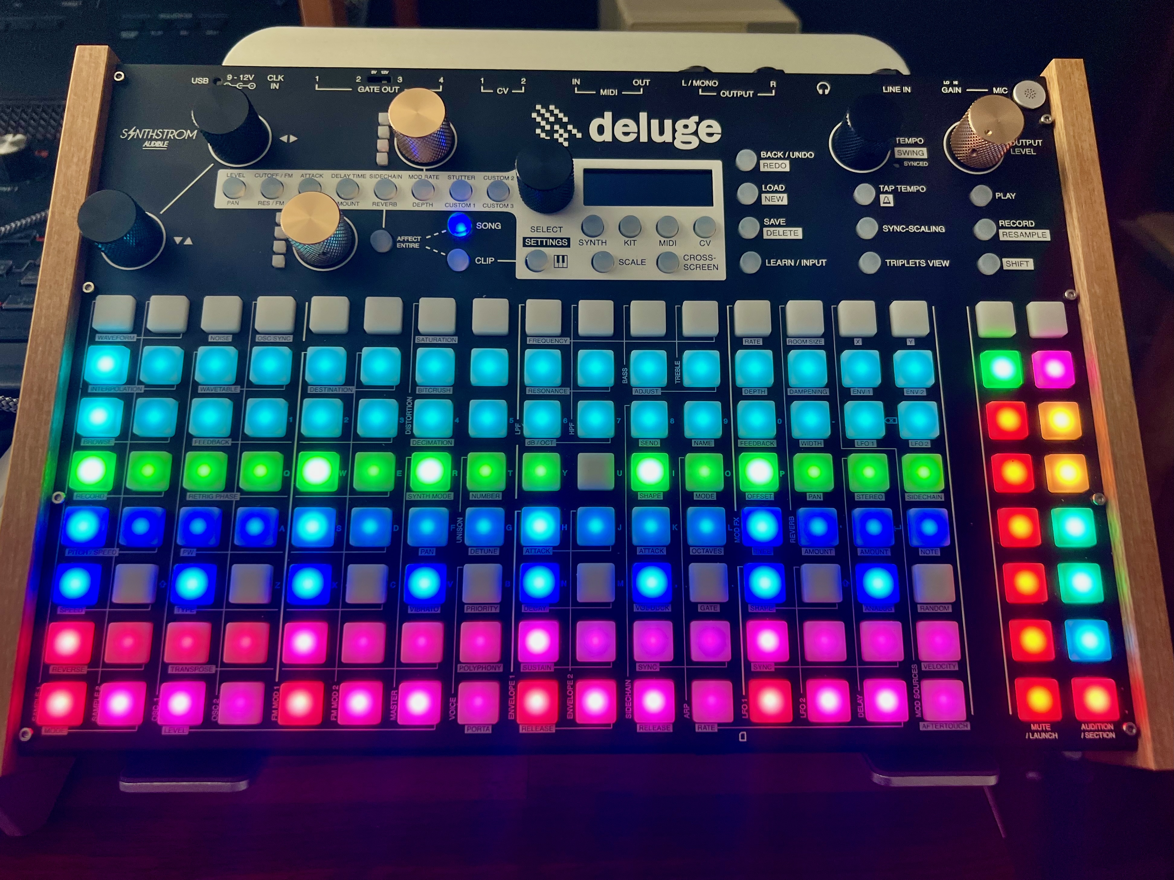 Synthstrom Deluge with 16x8 grid of RGB LED buttons plus two columns of buttons to the right for audition and mute; the upper section has knobs for controlling effects, tempo, volume, and navigation