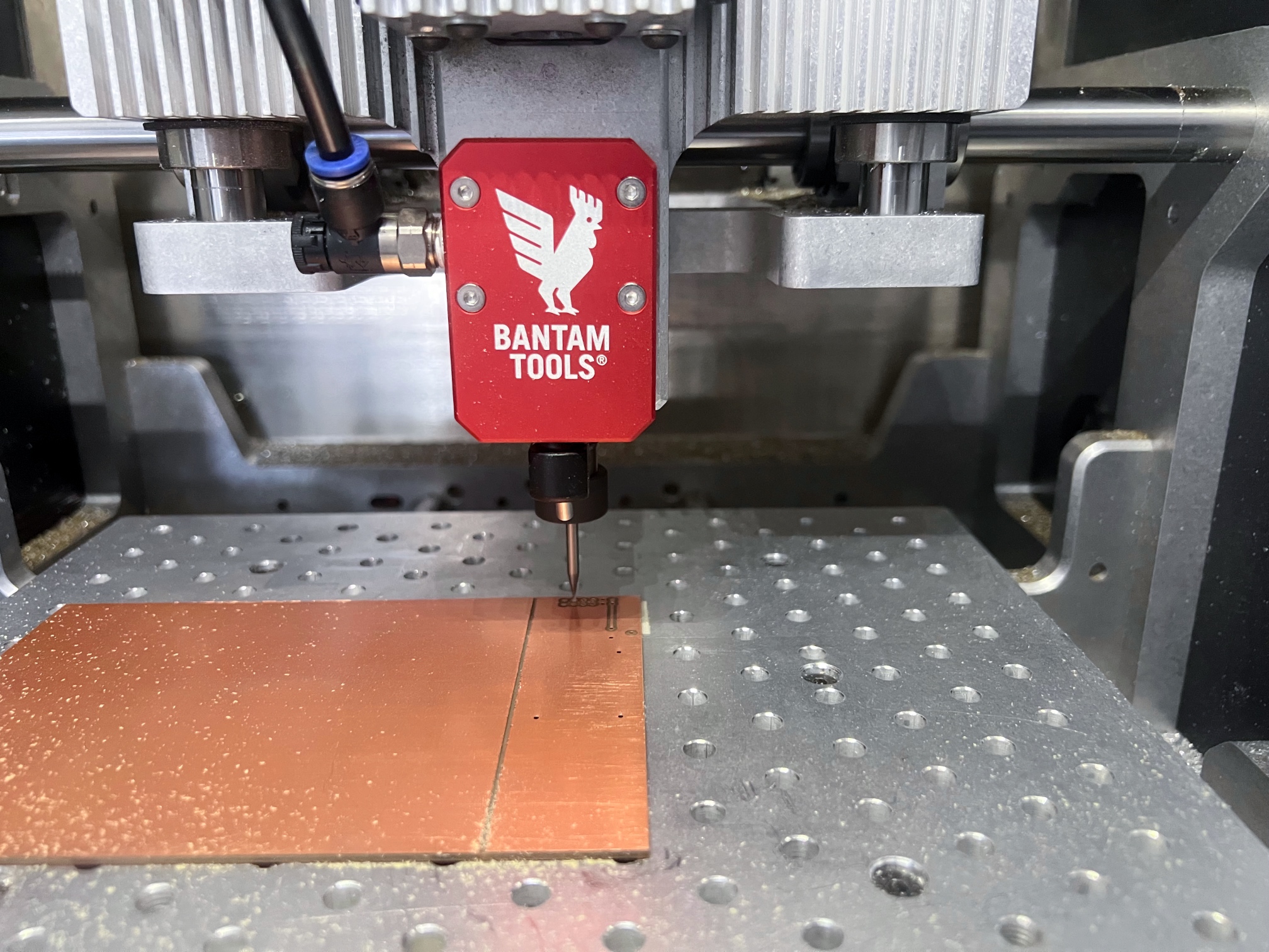 Bantam desktop CNC mill in action, isolation routing a test PCB in FR-1, with red anodized air-cooling nozzle adapter visible on front of the spindle above a fixturing pallet with regularly spaced threaded holes; PCB taped to the pallet