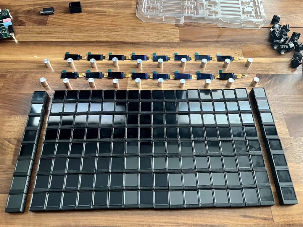 16x8 grid of 3D-printed key caps with LCDs assembled inside with an additional column on each side for controls and two rows of potentiometer knobs across the top with small, wide aspect ratio LCDs above each, all laid out on my desk surface